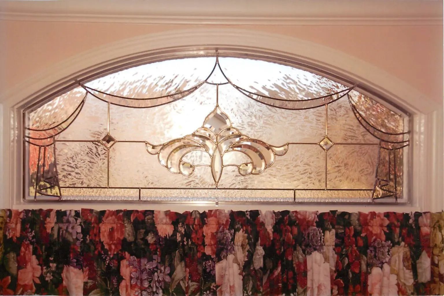 An arched stained glass window with a decorative floral design, above a floral-patterned curtain.