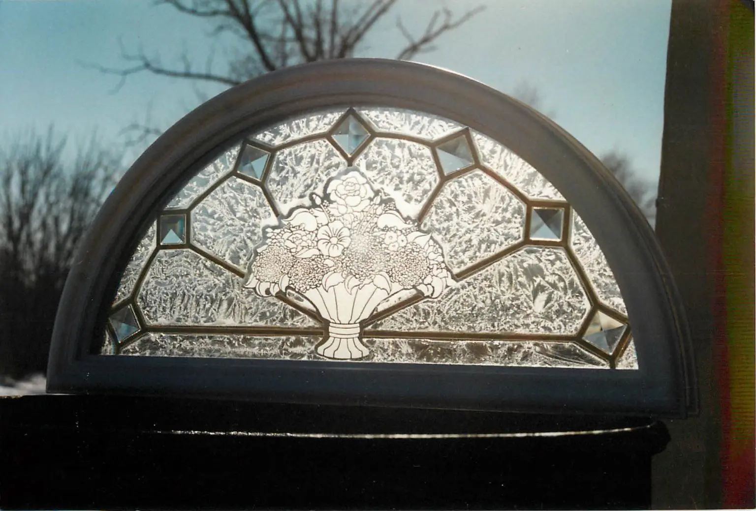 A semi-circular stained glass window with a floral basket pattern. The design features textured glass and geometric accents, set against an outdoor backdrop.