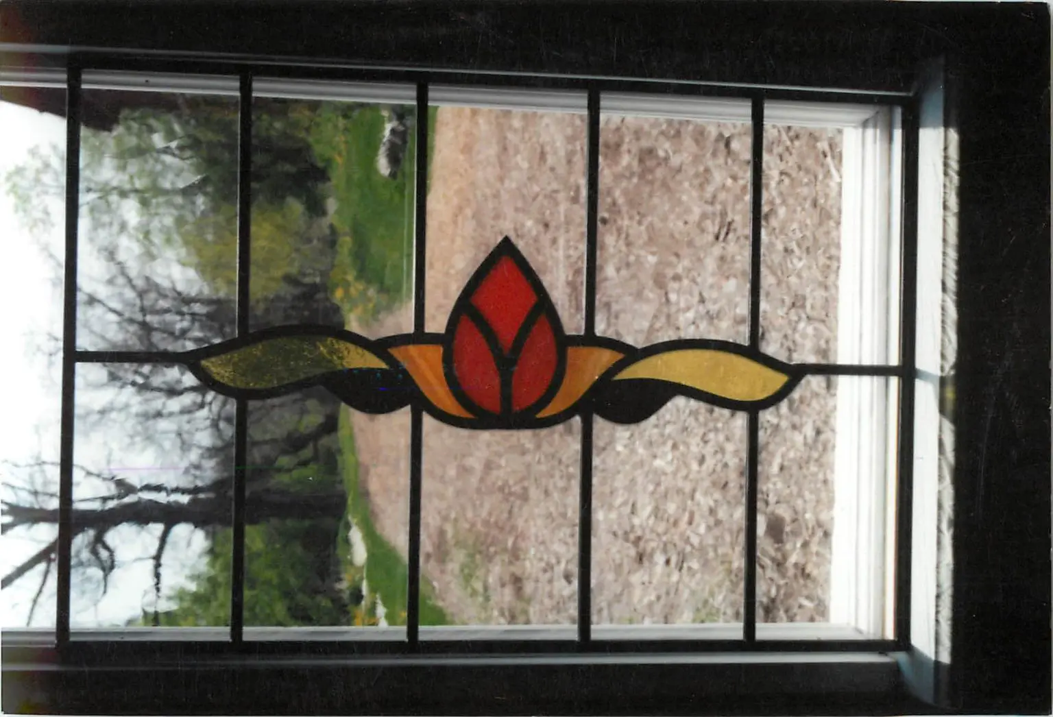 View through a window with stained glass featuring a red and yellow floral design; outside is a wooded area and lawn.
