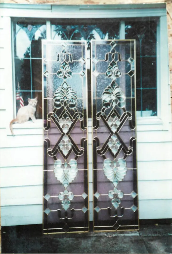 Two ornate glass doors with intricate designs are propped against the wall of a house. A cat sits on the windowsill in the background.
