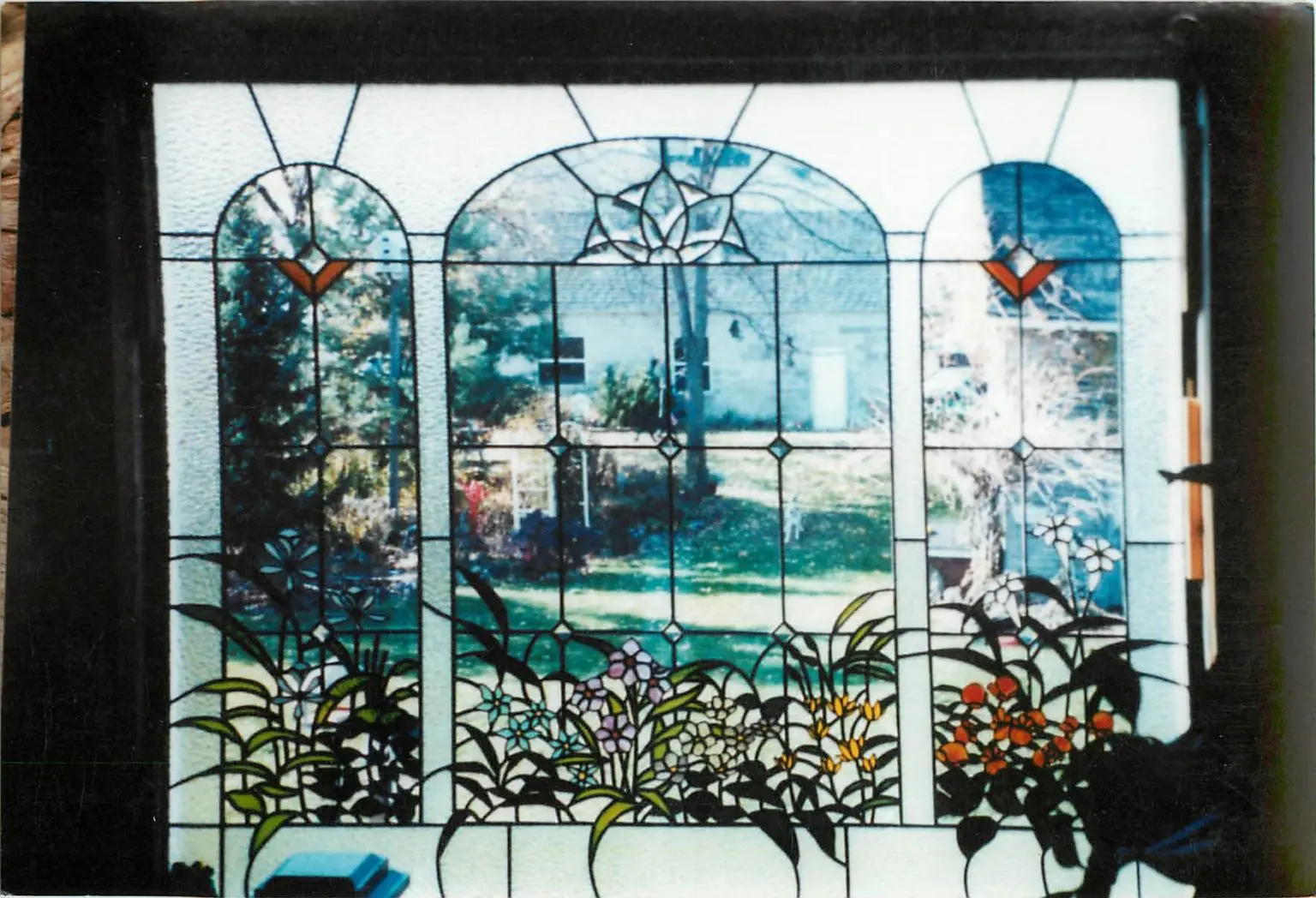 Stained glass window depicting various plants and flowers, with a view of a yard and house visible through the glass.