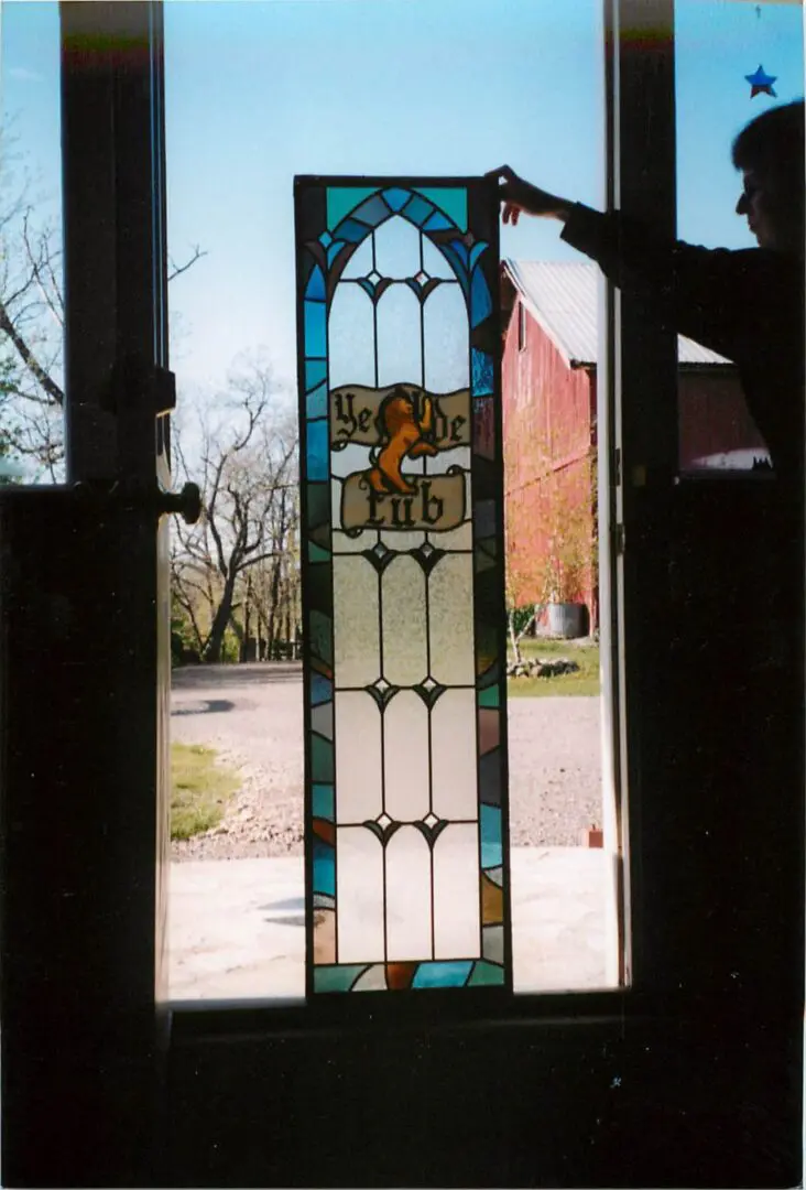 A person holds up a stained glass window panel with the words "Ye Olde Curio" and an orange lion emblem; a barn and trees are visible outside.