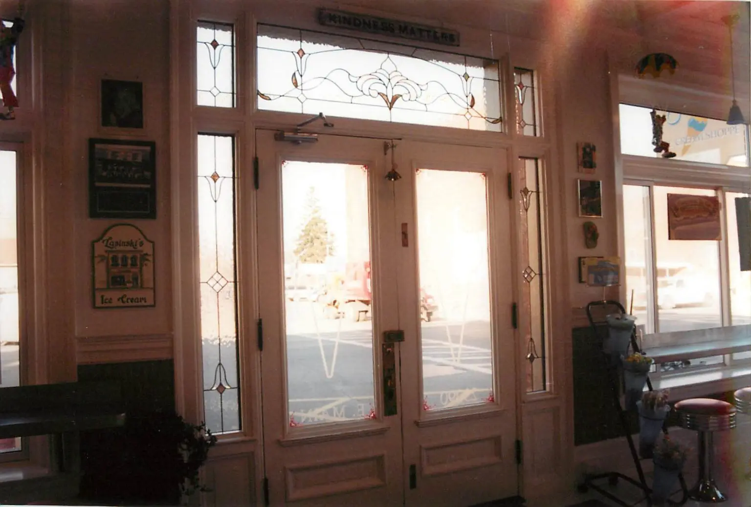 A decorative glass door with a "Kindness Matters" sign above it, flanked by framed pictures. Outside, a red truck is visible. Inside, there's an ice cream sign and a counter with items.