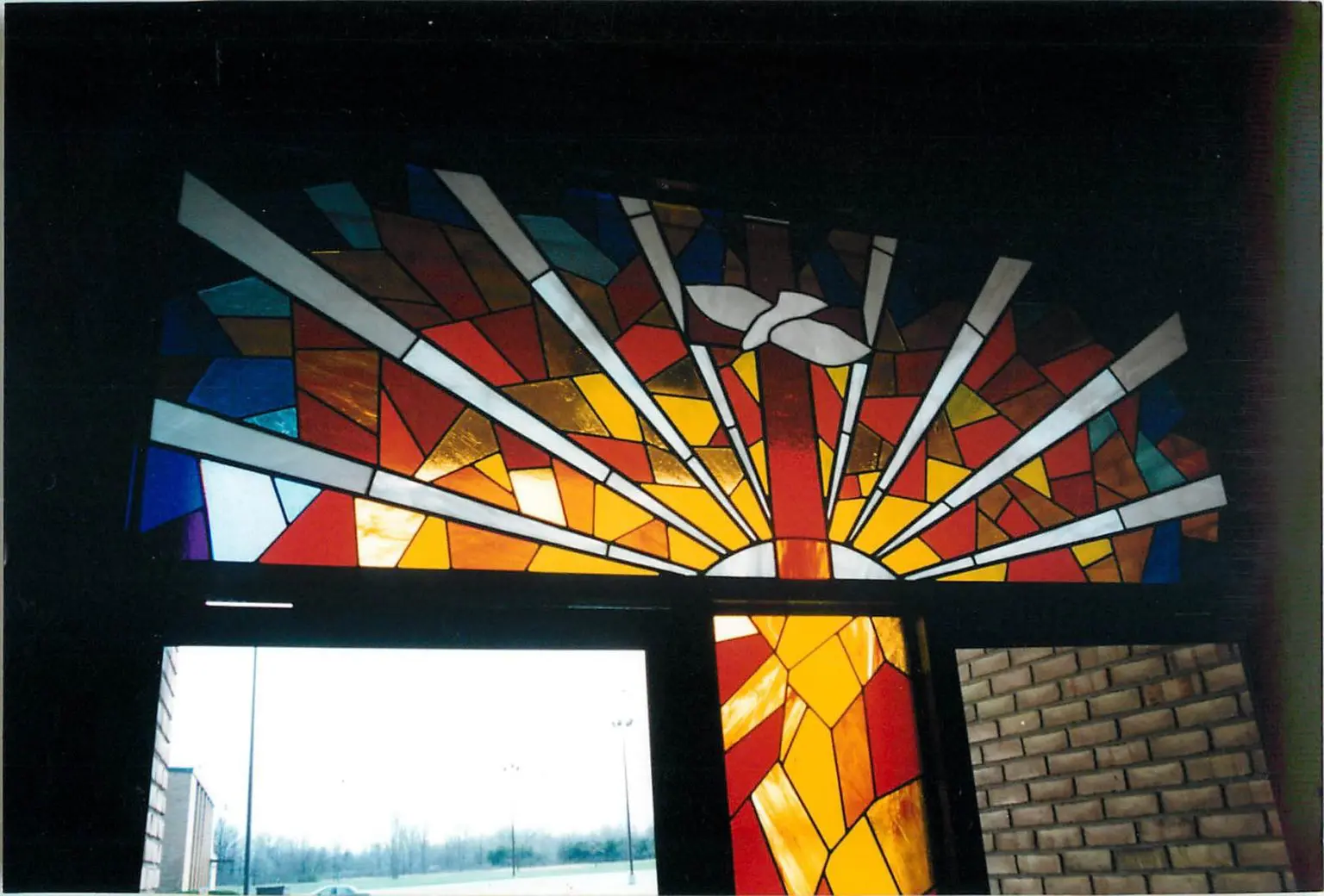 Stained glass window depicting a colorful sunrise with rays of light and a white dove in the center.
