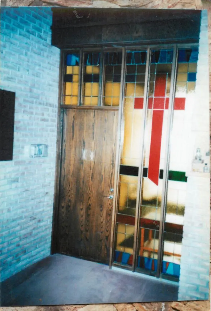 Wooden door beside stained glass panels featuring a red cross and multicolored designs in a brick-walled room.