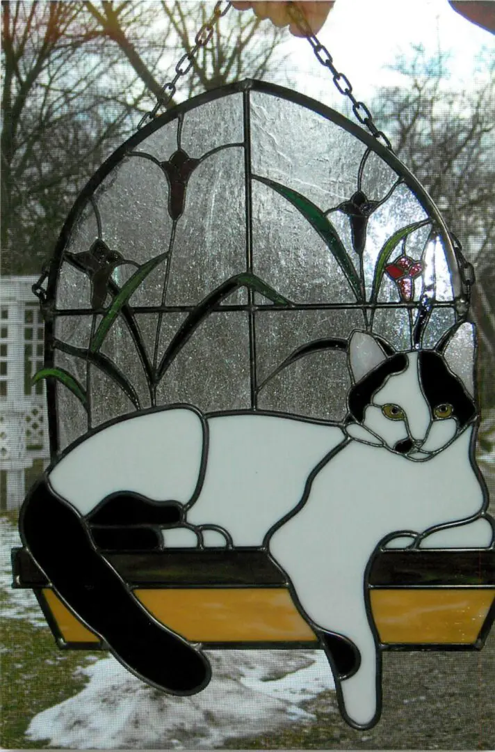 Stained glass art of a black and white cat lounging on a swing against a garden background.