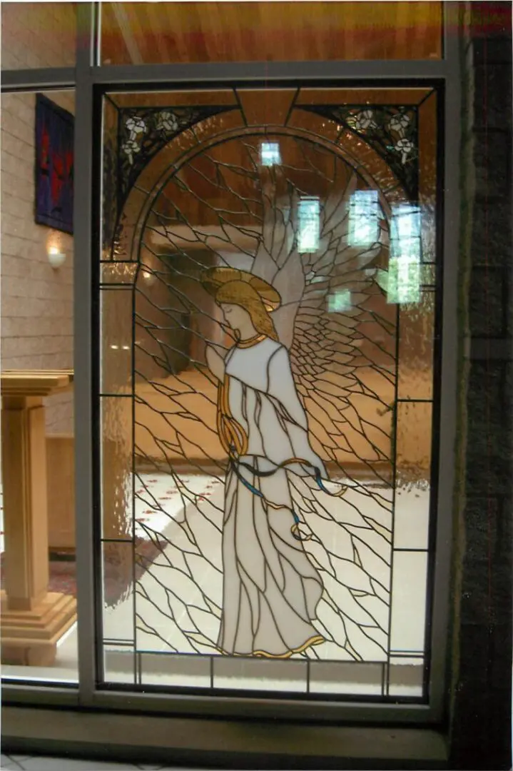 Stained glass window depicting an angel in white robes with large wings, set in a decorative arch frame.