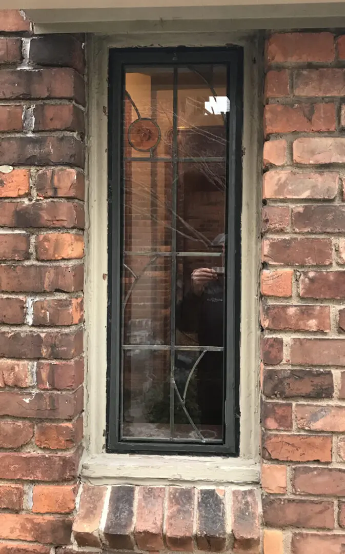 Tall, narrow window set in a brick wall. A person is faintly reflected, holding a phone. A small circular stained glass piece is at the top.