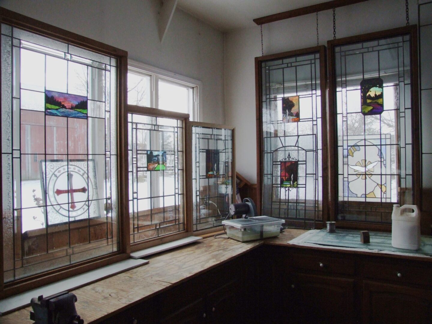 Stained glass panels with colorful designs are displayed in a sunlit room. A wooden countertop is below the windows, holding a few items, including a small bucket and a white container.
