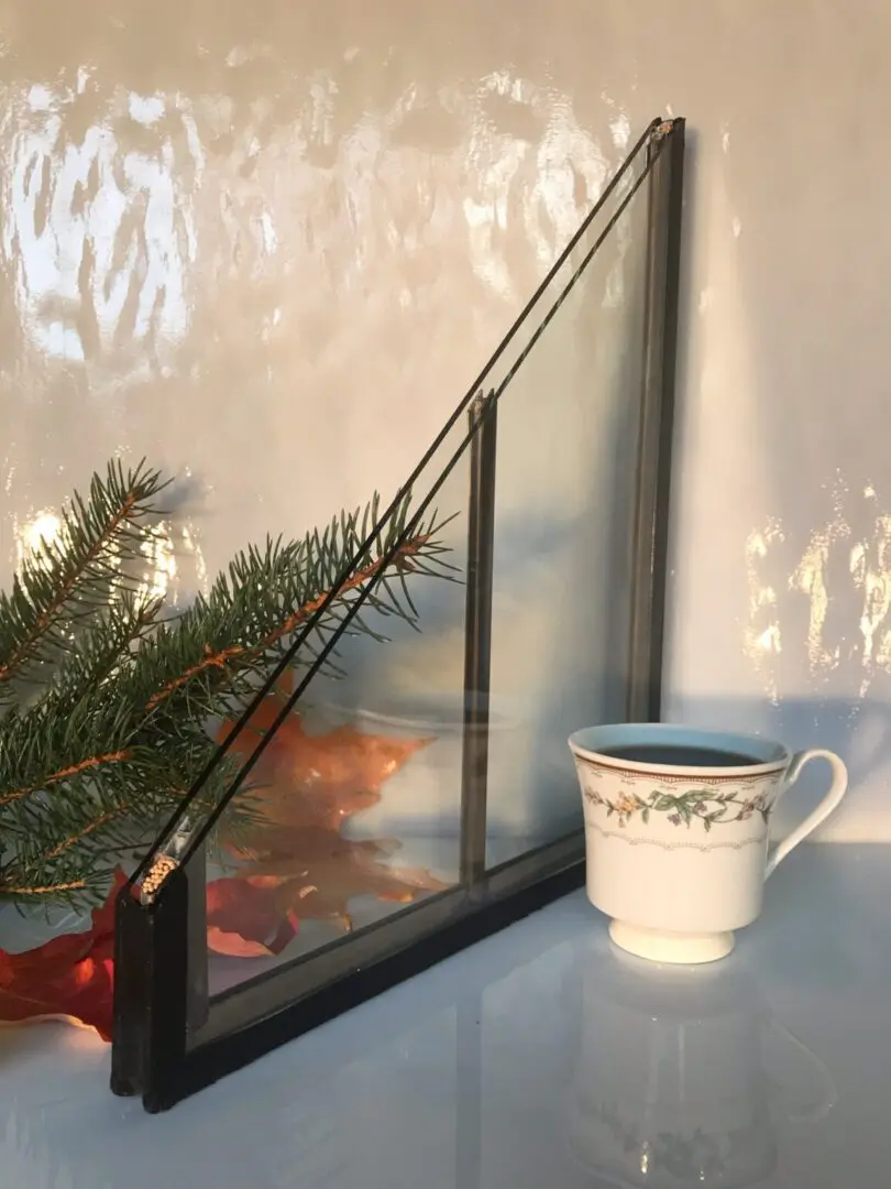 Glass window corner sample with a black frame sits next to a white cup decorated with floral patterns. Pine branches and autumn leaves are in the background.
