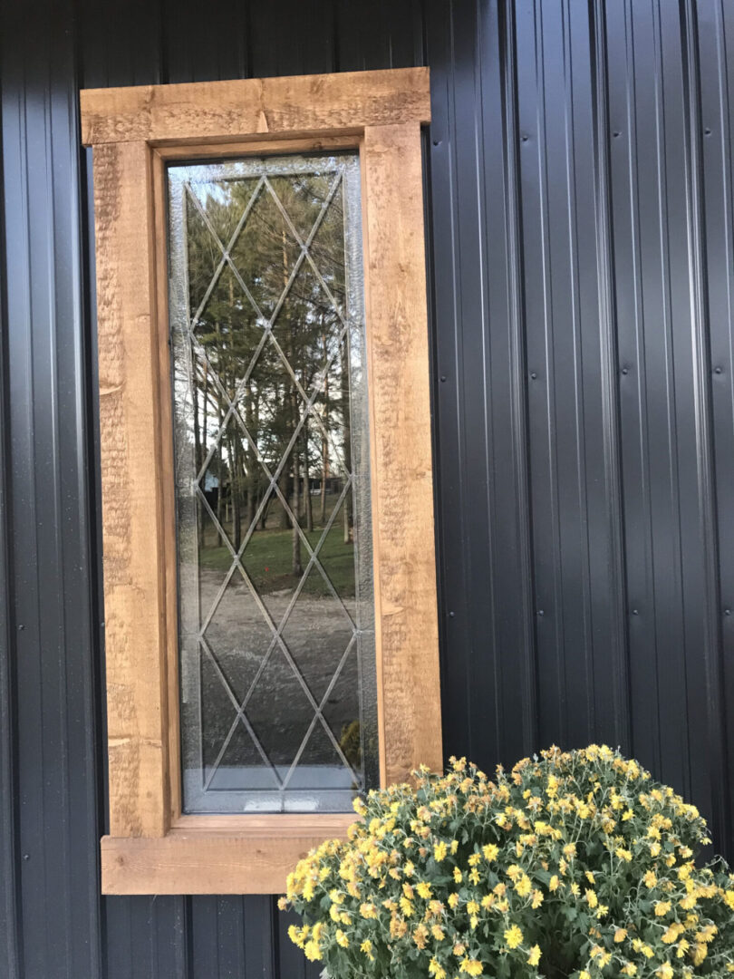 Tall window with wooden frame and diamond grid design on a dark paneled wall. Yellow flowers in a pot below. Trees are reflected in the window glass.