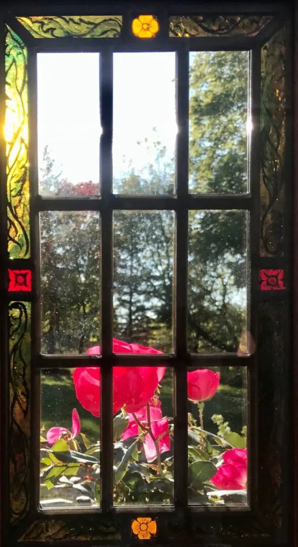 Stained glass window with a floral border frames a view of red roses and green trees under a blue sky.