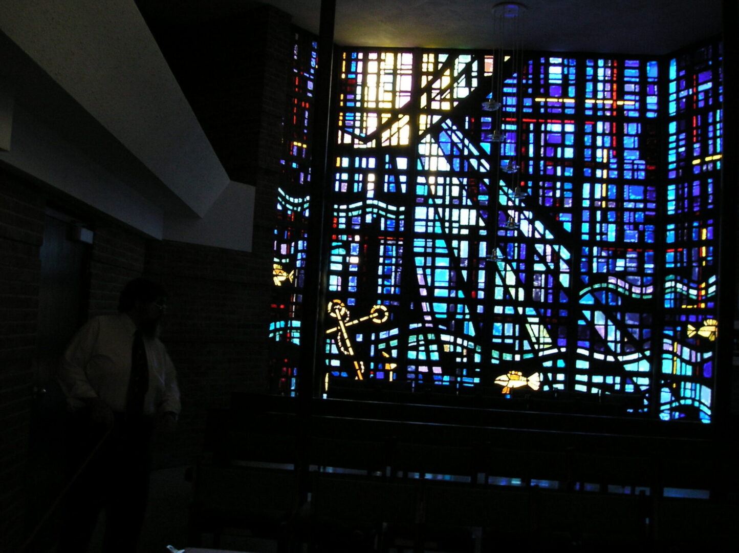 Dimly lit room with a large, colorful stained glass window featuring abstract patterns. A person stands near the left side, partially shadowed.