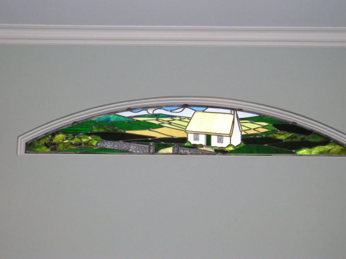 Arched stained glass window depicting a house and green landscape scene, set in a light gray wall.