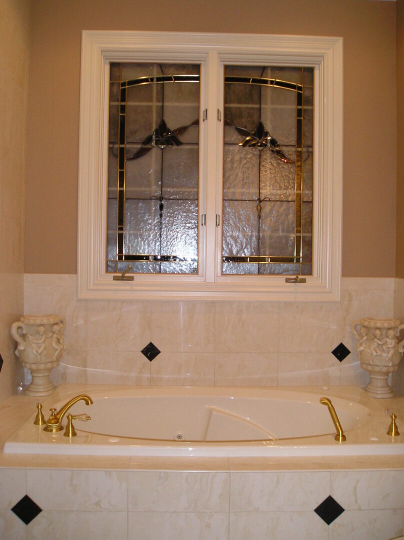 A luxury bathroom featuring a white bathtub with gold fixtures, set against a tiled wall. Above the tub, a stained glass window is framed by beige walls. Two decorative urns flank the tub.