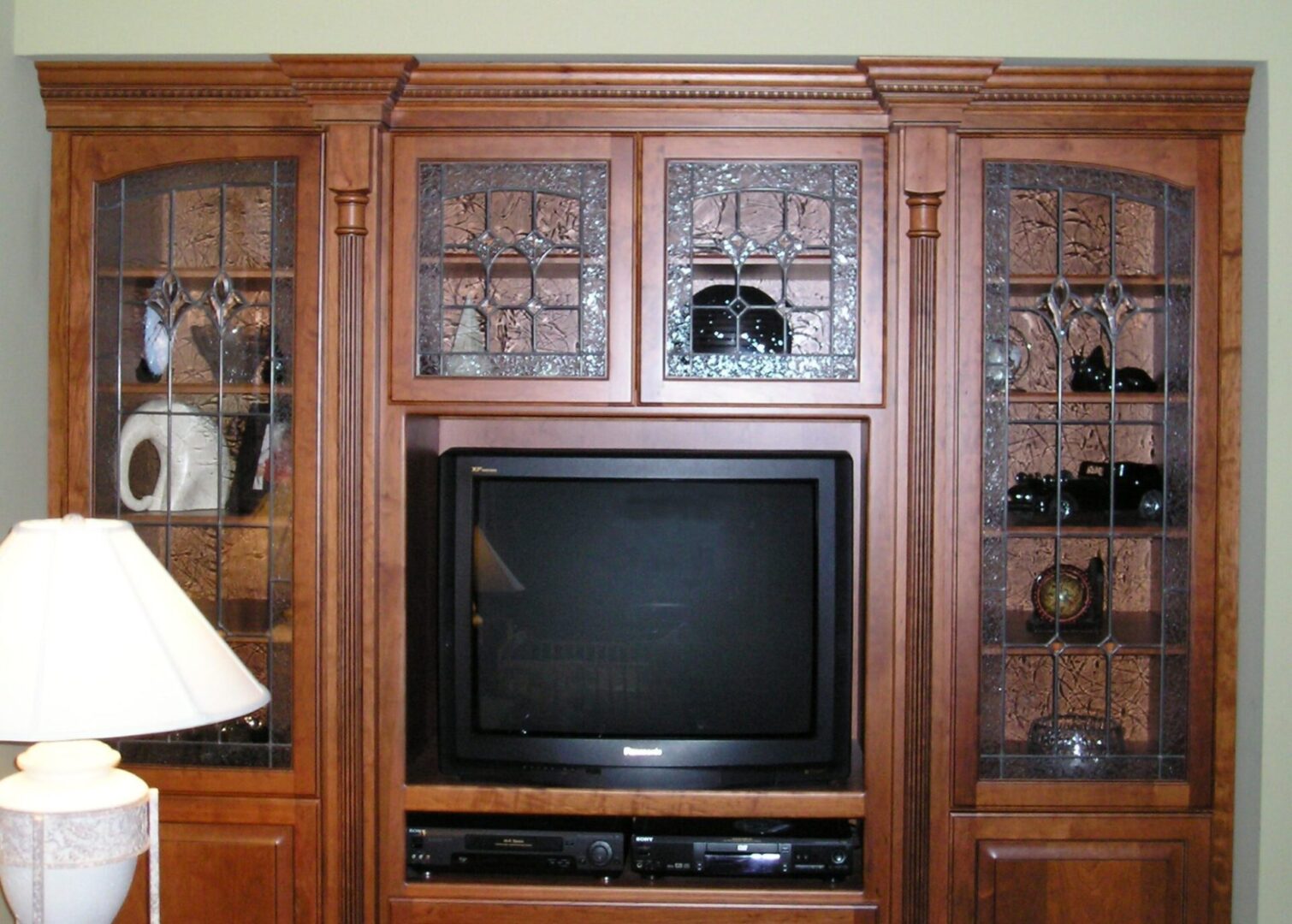 Wooden entertainment center with a TV in the middle, flanked by cabinets with glass paneling displaying decorative items. A white lamp is partially visible on the left.