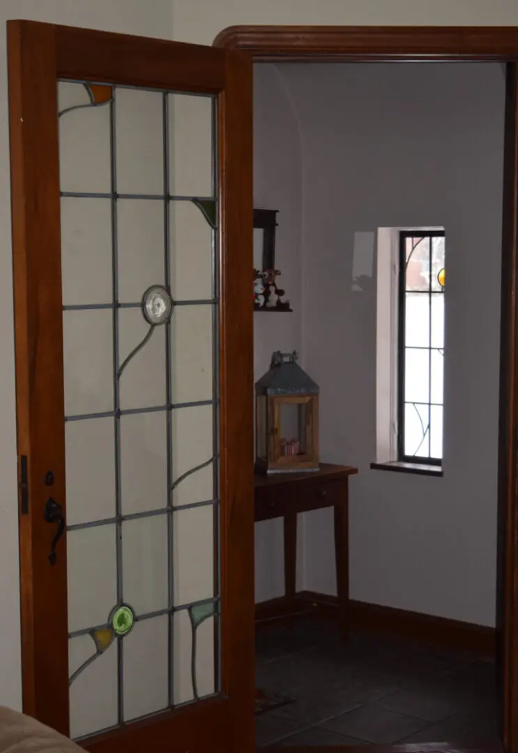 Wooden door with a stained glass panel, partially open, leading to a room with a small table, a lantern, and a small window in the background.