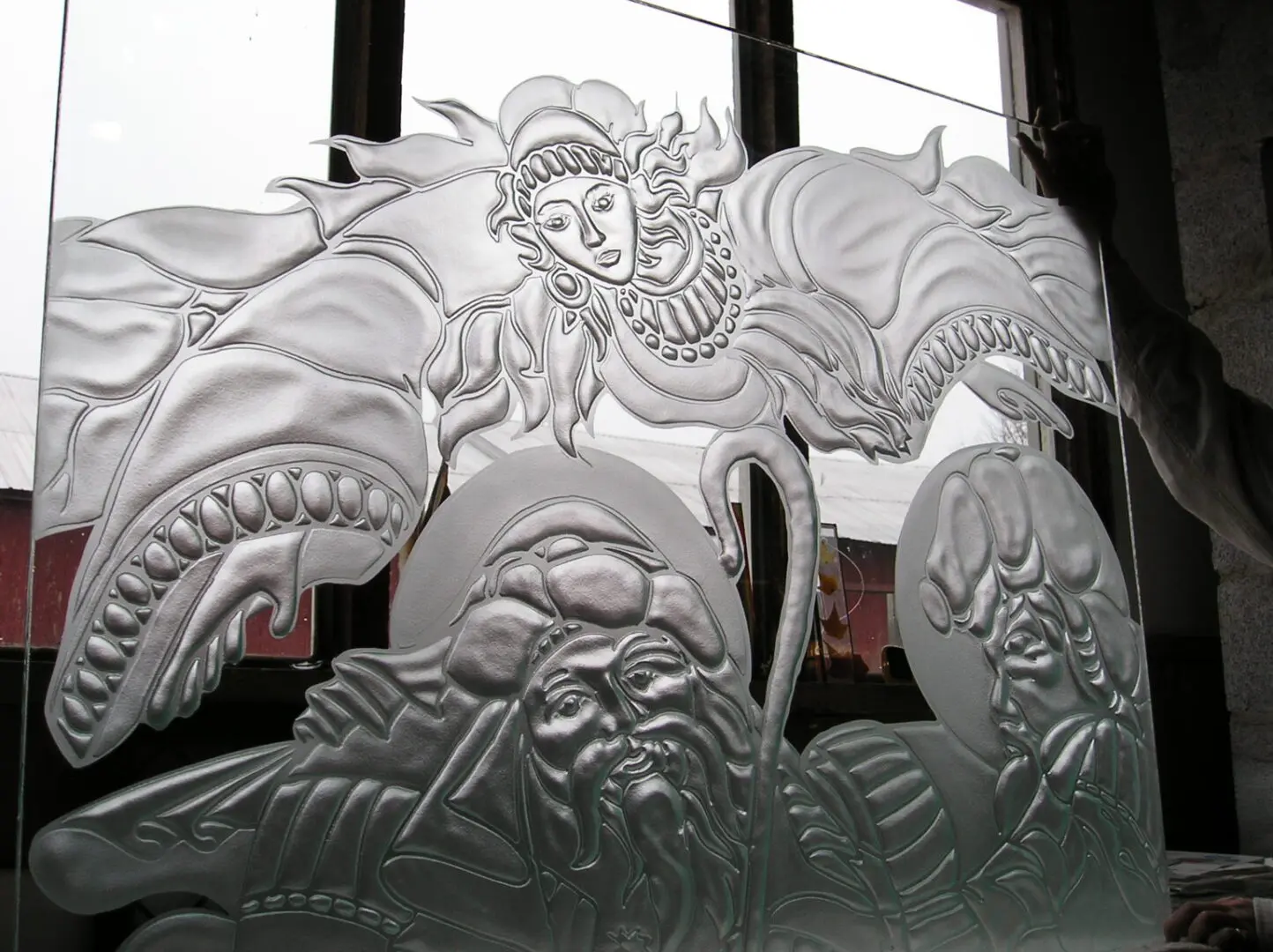 Intricately carved glass panel depicting a mythical scene with a dragon and three figures. Background shows a window with natural light entering.