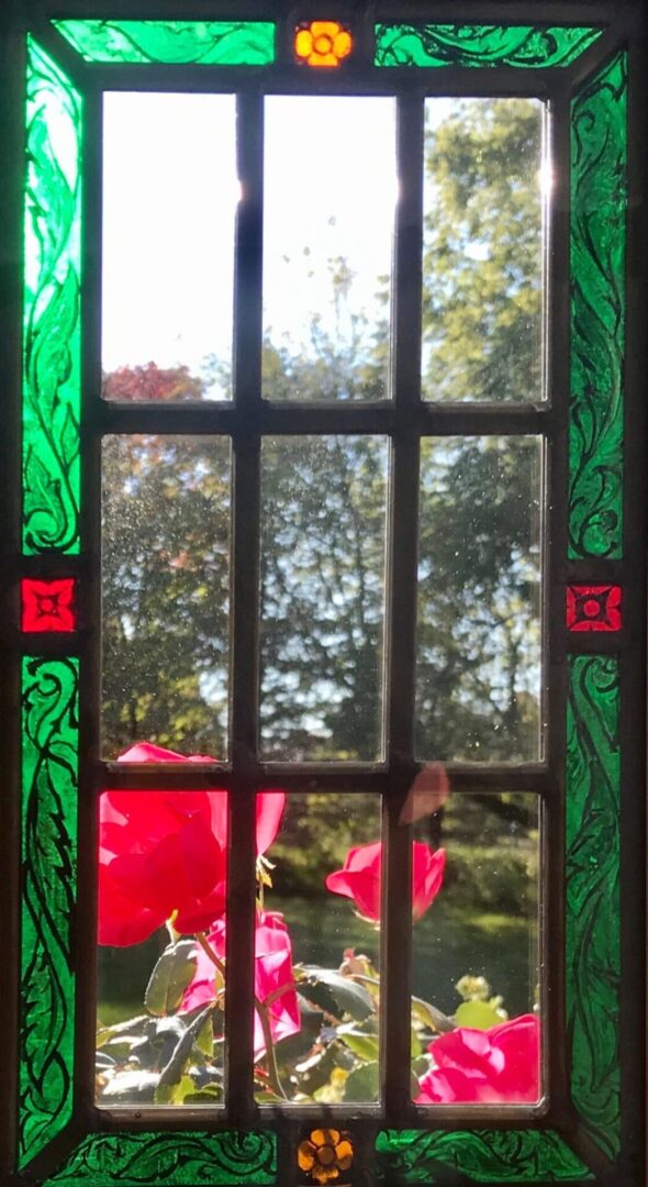 Stained glass window with a colorful green and red border. Outside, red roses and a blurred view of trees are visible in the sunlight.